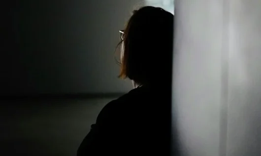 almost black and white photo of a woman with a bob (the ends of her hair are brown) and glasses sitting with her back to the viewer and her back against a wall on the right hand side, with a window at the top right hand side. The light of her round digital watch is illuminated.