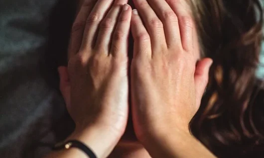 guilt_and_shame_jannes-jacobs-1zoWx-Cikb8-unsplash Photo of a woman lying on a bed (head, chest, forearms and hands), with the sheets and pillow behind her, with hands covering her face, with long brown hair falling behind her head, with a low-cut grey unsleeved top.