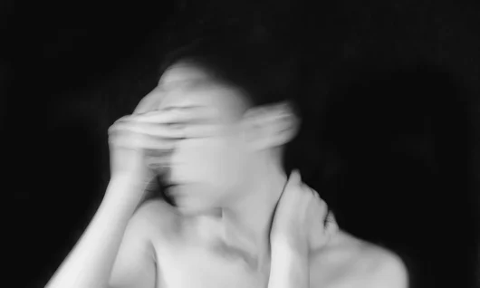 menopause_anna-stampfli-pKaK2FiN2IE-unsplash Blurred black and white photo of a woman seemingly shaking her head (with her head looking towards the left) with her hand over her face with her other hand on her neck covering her bare shoulders, with a black background.
