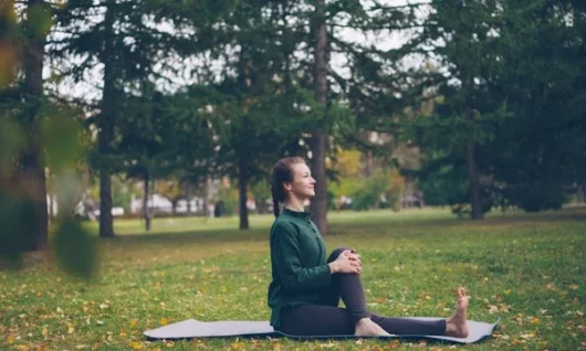 polyvagal_theory_vitaly-gariev-ub6cttunrrw-unsplash Young smiling woman looking forward (to the right) with a platted pony tail and bare foot, in a green long-sleeved top and long black leggings, sat with one leg outstretched and the other bent over the first and her hands clasped around the bent knee on a yoga mat, in a park with grass, some blurred buildings and trees behind her.