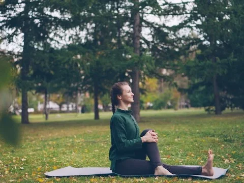 Young smiling woman looking forward (to the right) with a platted pony tail and bare foot, in a green long-sleeved top and long black leggings, sat with one leg outstretched and the other bent over the first and her hands clasped around the bent knee on a yoga mat, in a park with grass, some blurred buildings and trees behind her.